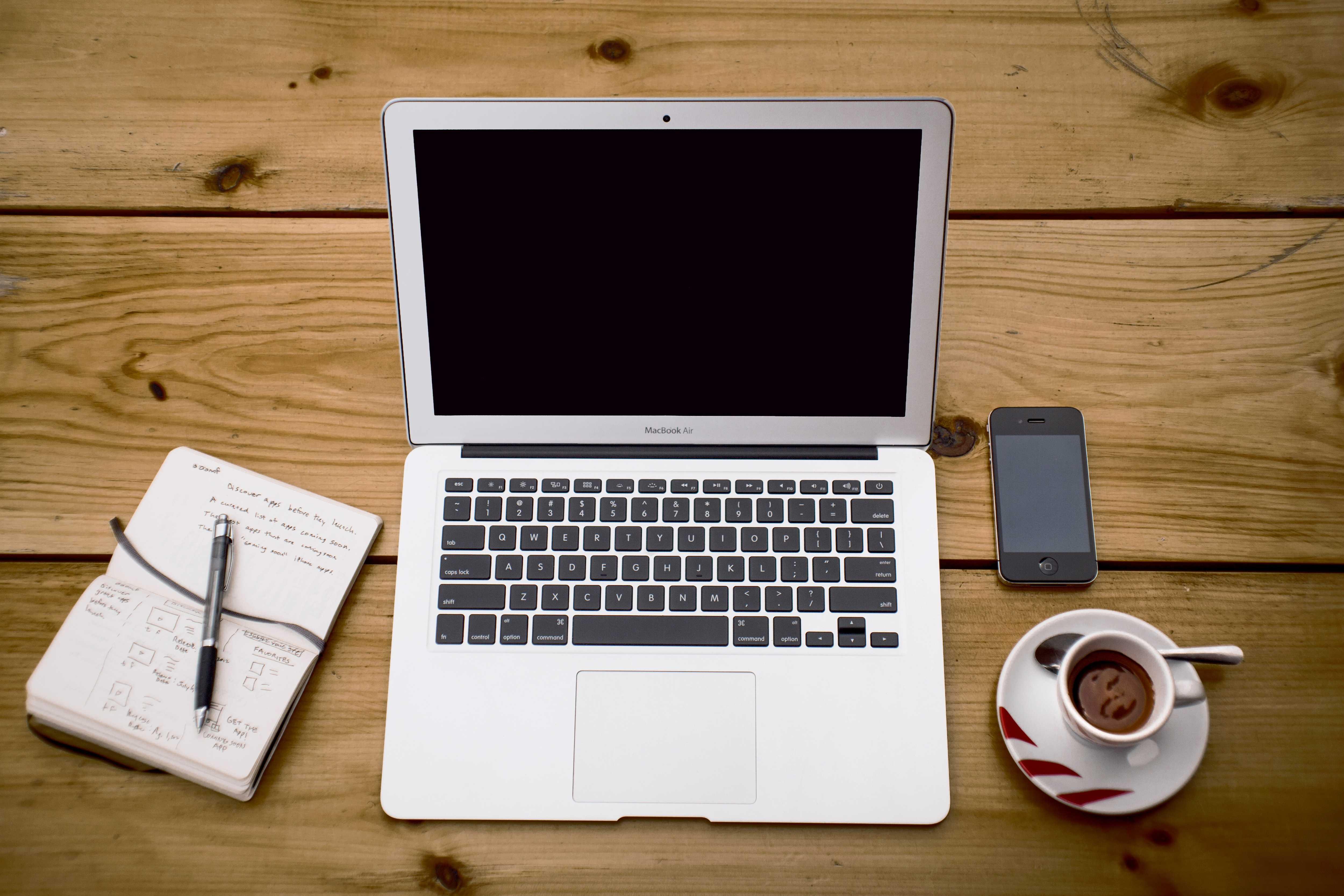 A 16:9 image of a macbook in the middle of a wooden table, an open notebook and pen on the left and an iPhone 3GS on the right with a cup of espresso located underneath it